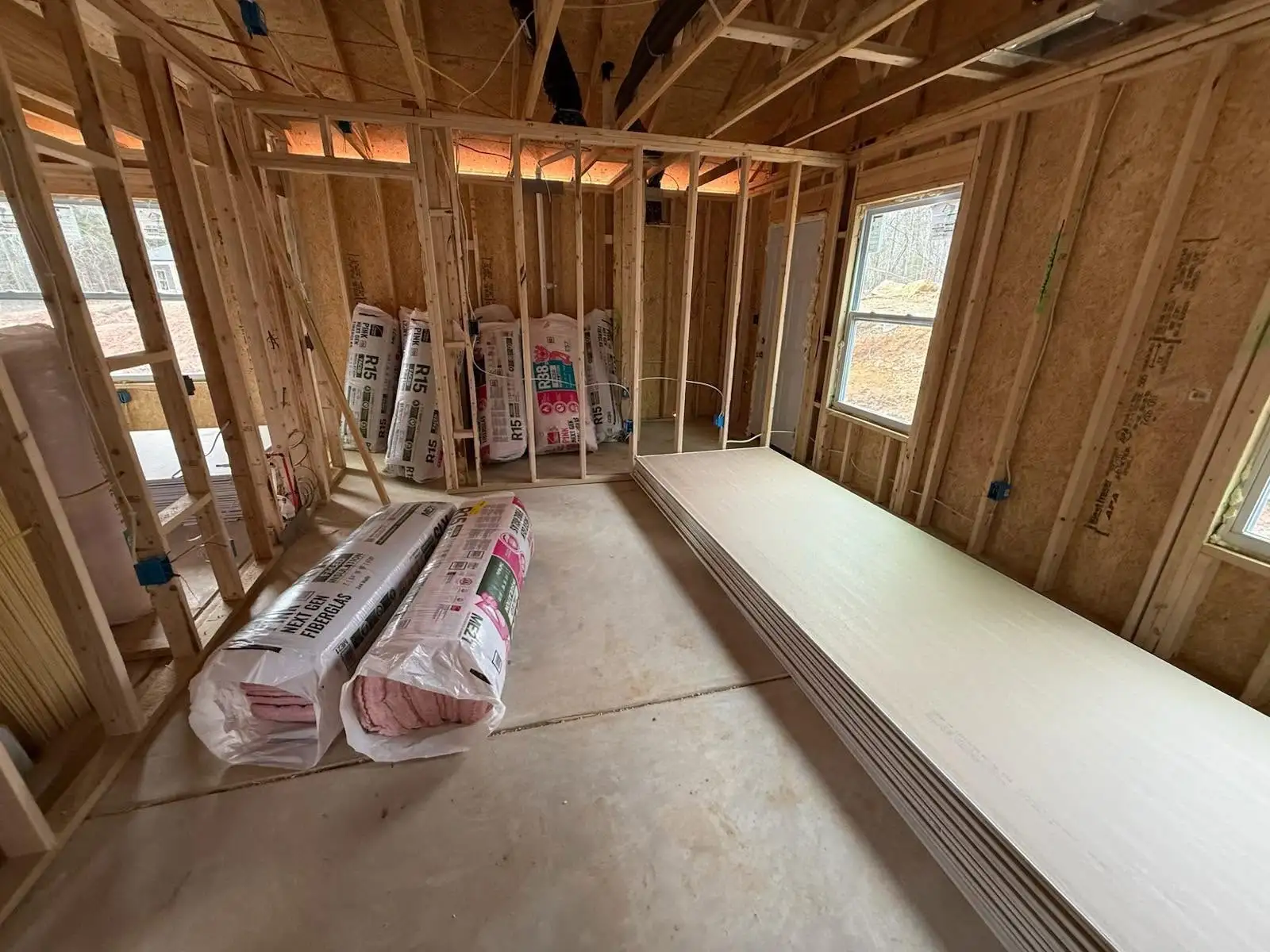 A framed room with stacked drywall sheets on the floor and bags of insulation standing against the framing, with windows letting in daylight.