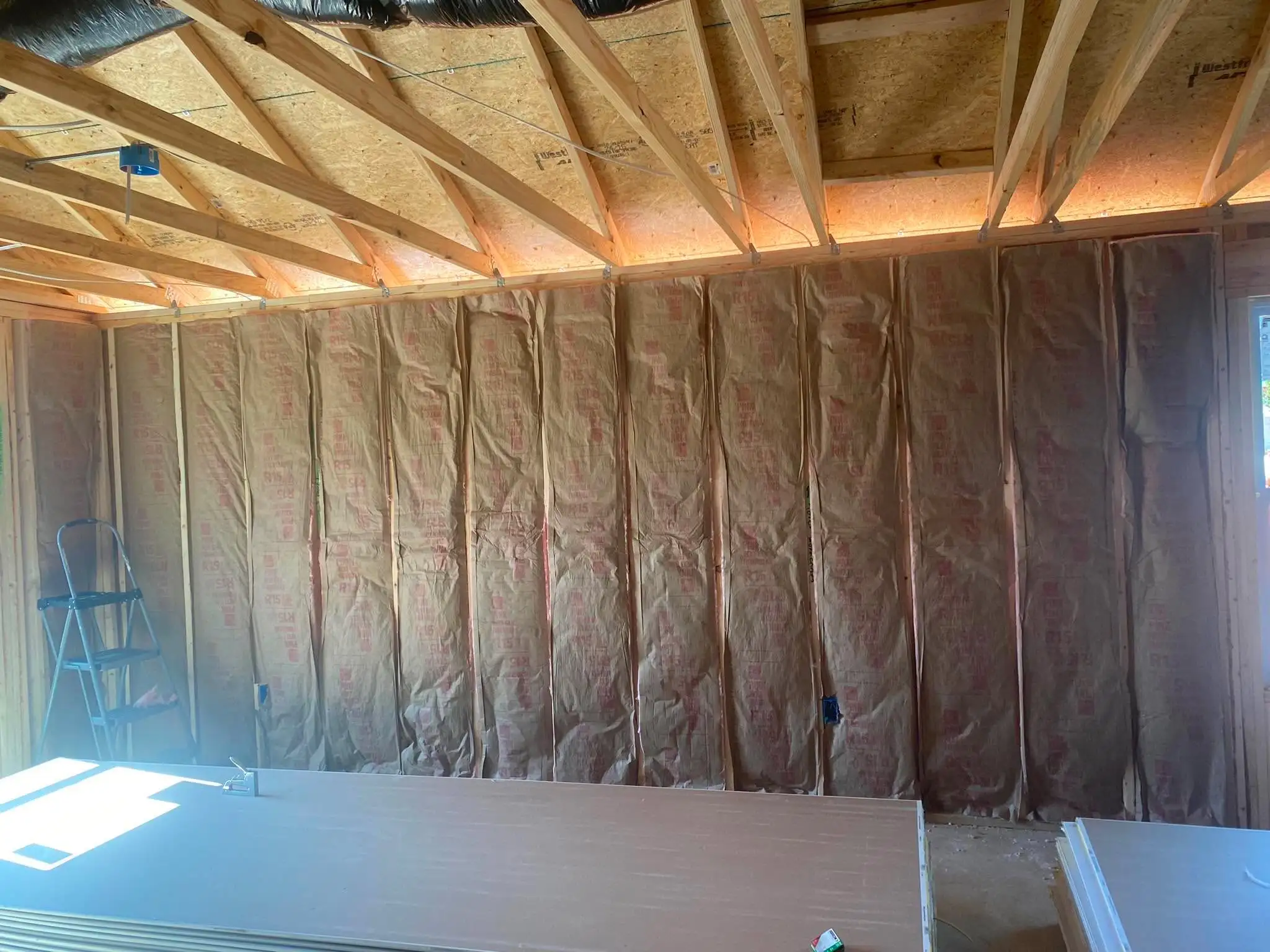 A framed wall filled with paper-faced insulation under exposed roof trusses, with a stepladder at the left and a stack of drywall sheets in front.