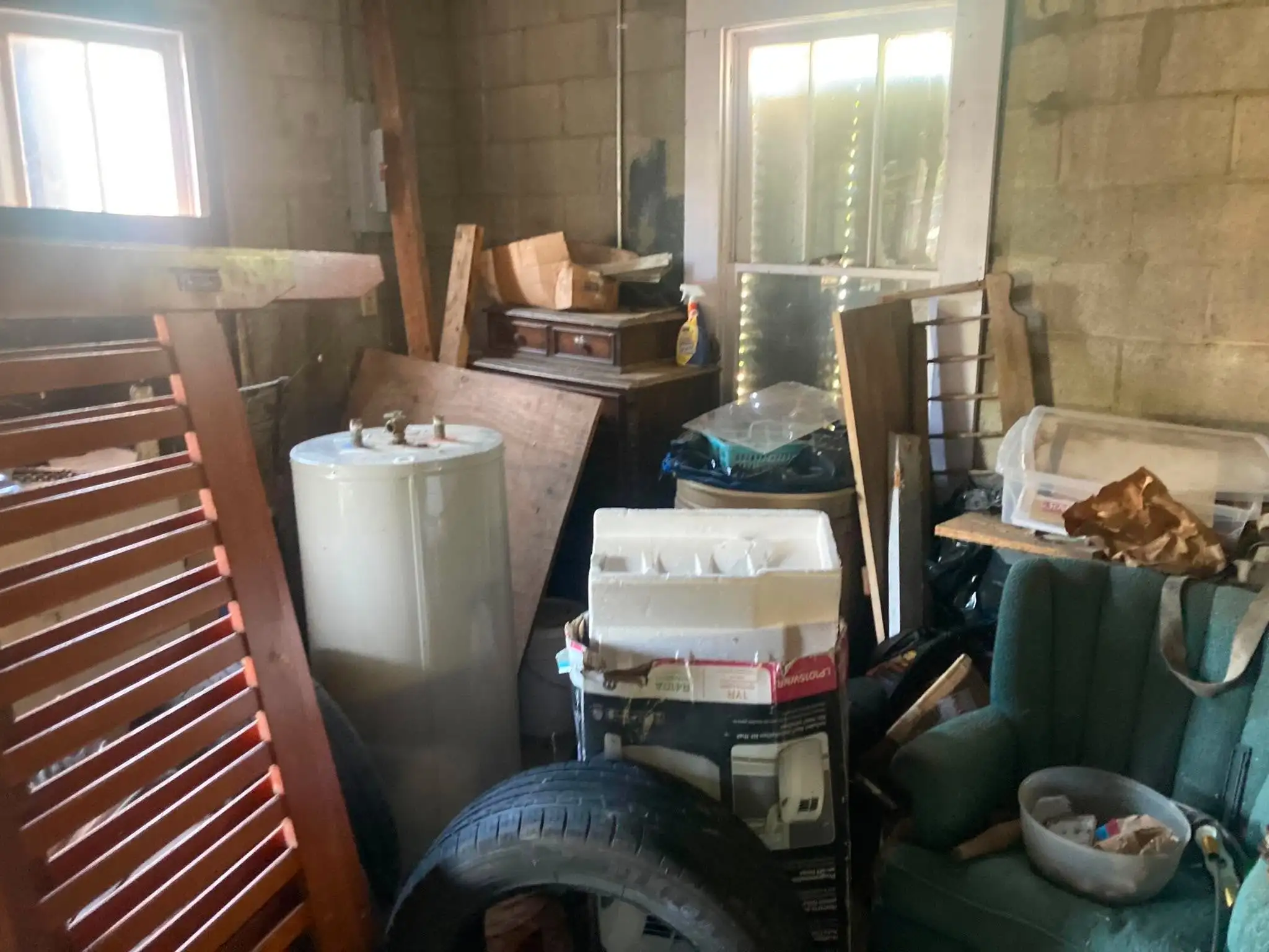 A garage packed with an old water heater, tires, a slatted wooden panel, boxes and an armchair against concrete block walls.