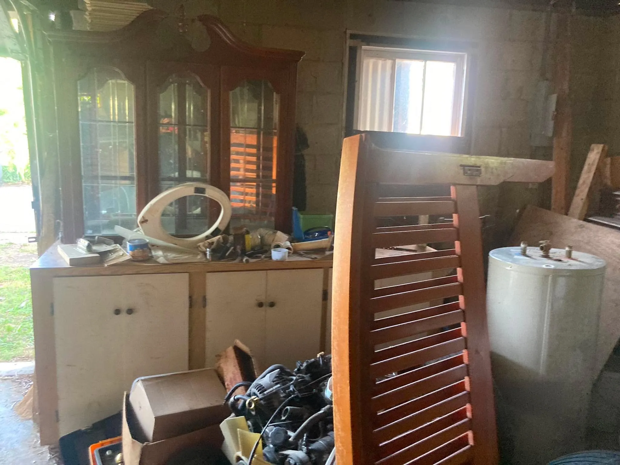A concrete block storage room crowded with an old wooden china cabinet, cabinets, and a water heater.
