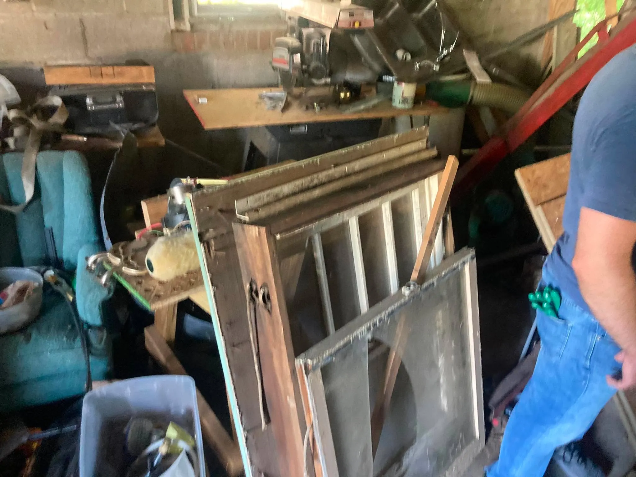 A cluttered workshop interior with sheet-metal panels, hand tools, and an old bench seat, with a person standing among the clutter.