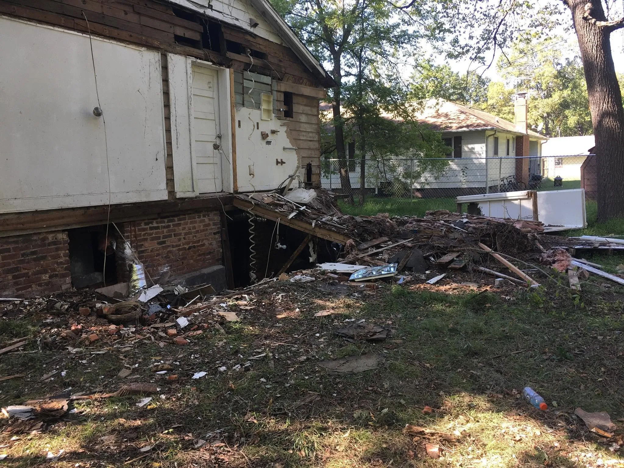 Another angle of the same exterior wall opening and debris pile, with a chain-link fence and neighboring house in the background.