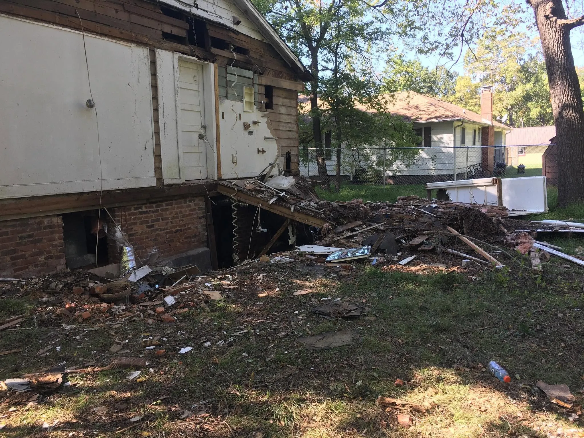The rear exterior of a house with siding removed and the brick foundation exposed, with a large pile of demolition debris in the yard below.