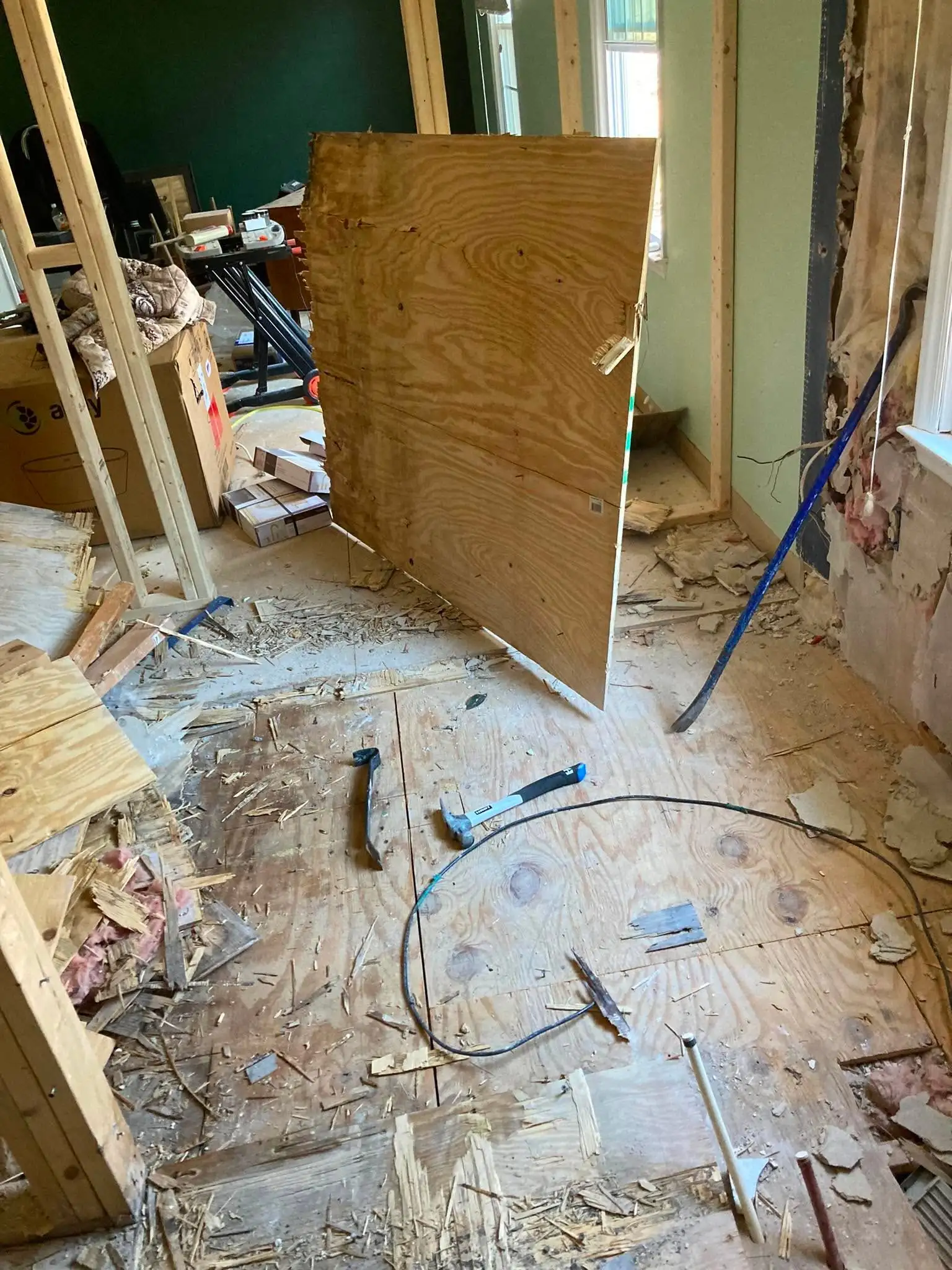 An interior room mid-demolition with a sheet of plywood stood upright, wall studs exposed, and a pry bar, hammer and debris on the floor.