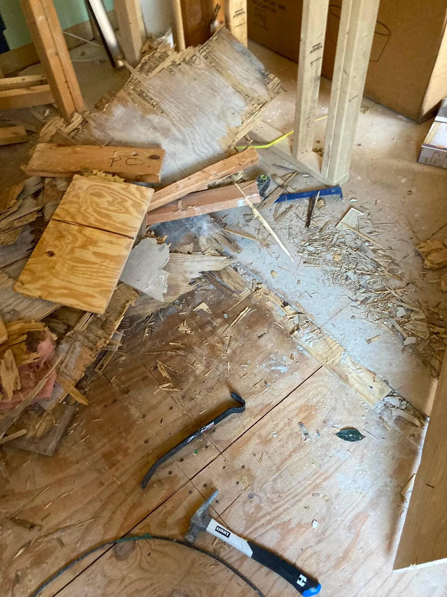 A torn-up plywood subfloor with broken boards piled around a pry bar and hammer.