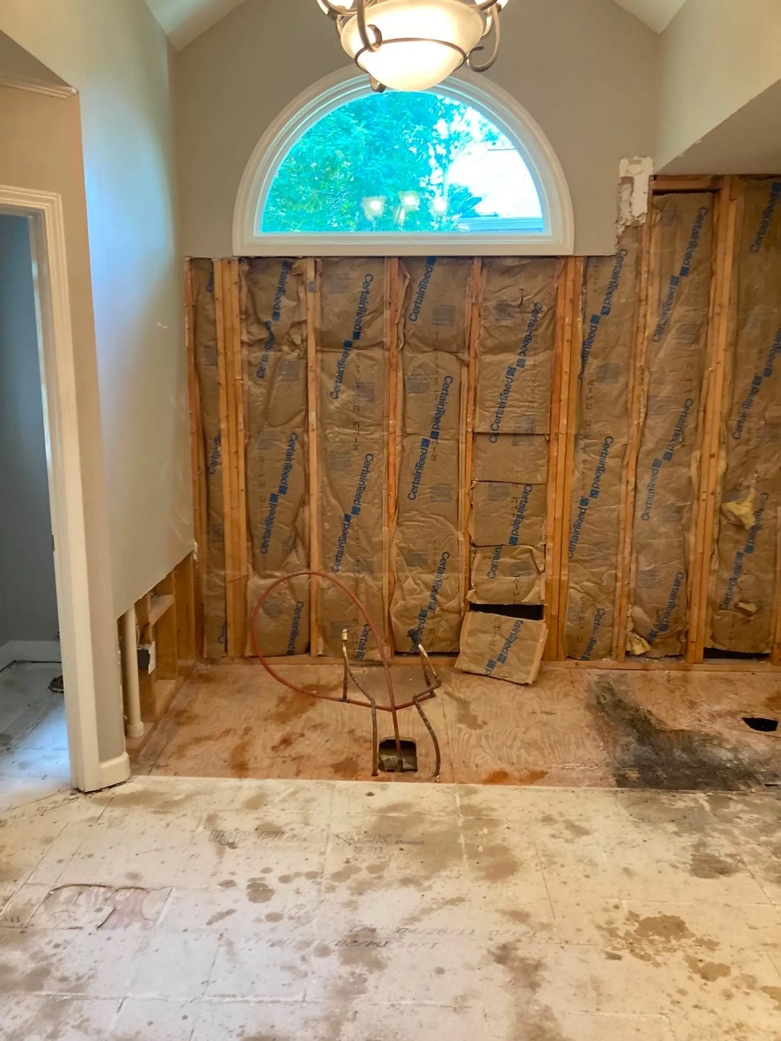 A room with a half-round arched window and chandelier, its lower wall stripped down to exposed studs and insulation.