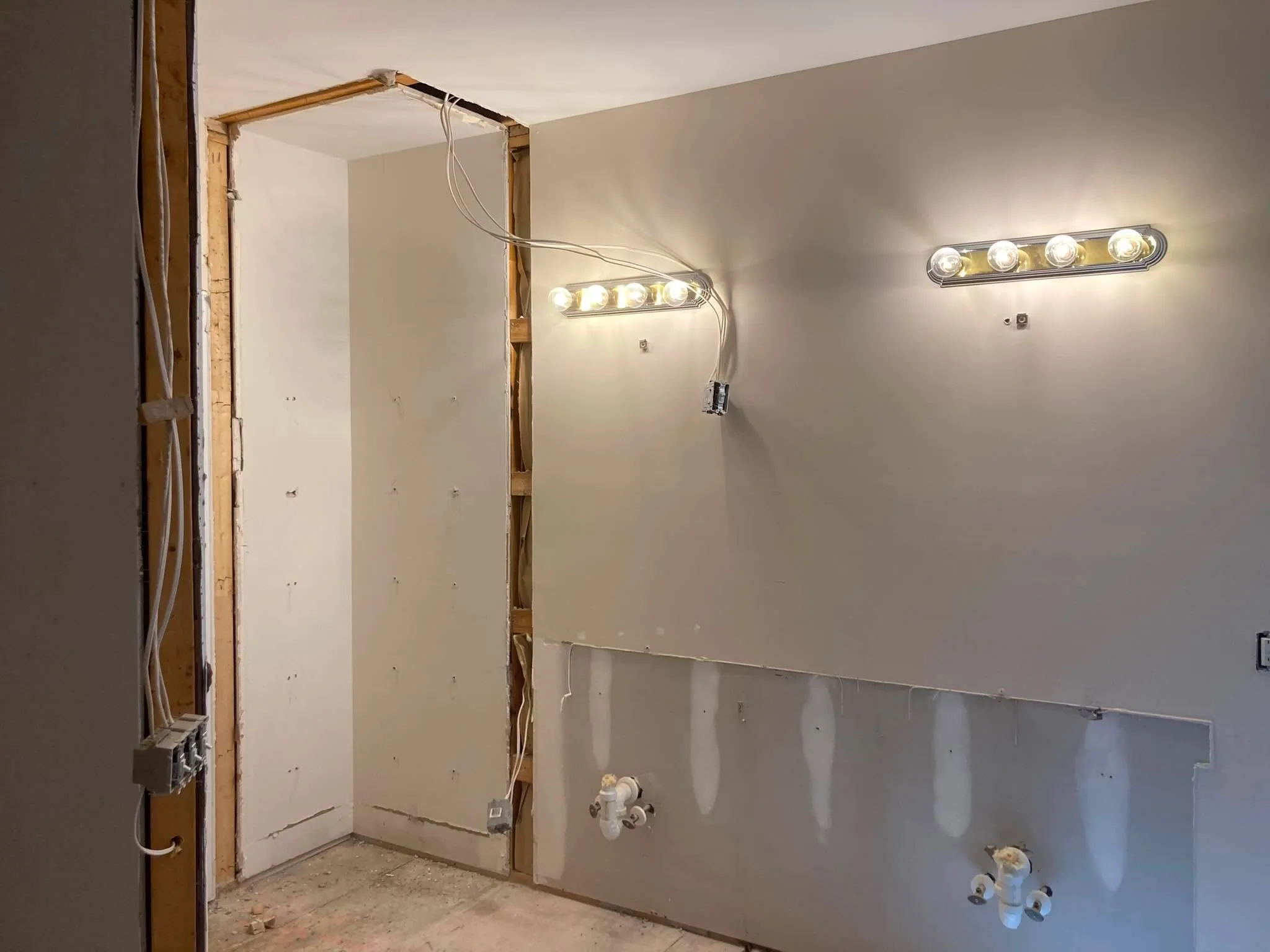 A room with two new vanity light fixtures mounted above a section of wall opened down to the studs and plumbing stub-outs.