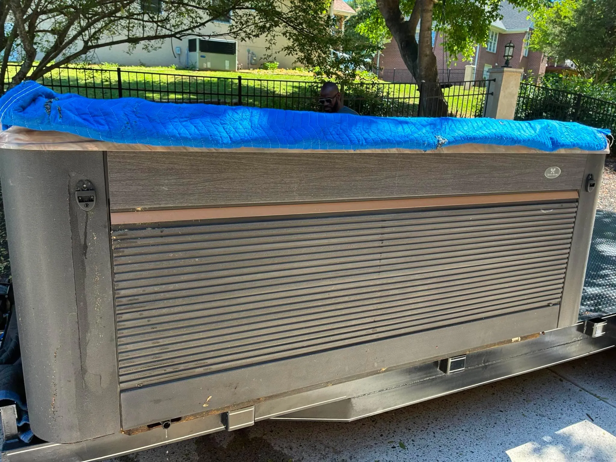 A hot tub lying on its side with a slatted skirt and a blue cover, loaded for transport beside an iron fence.